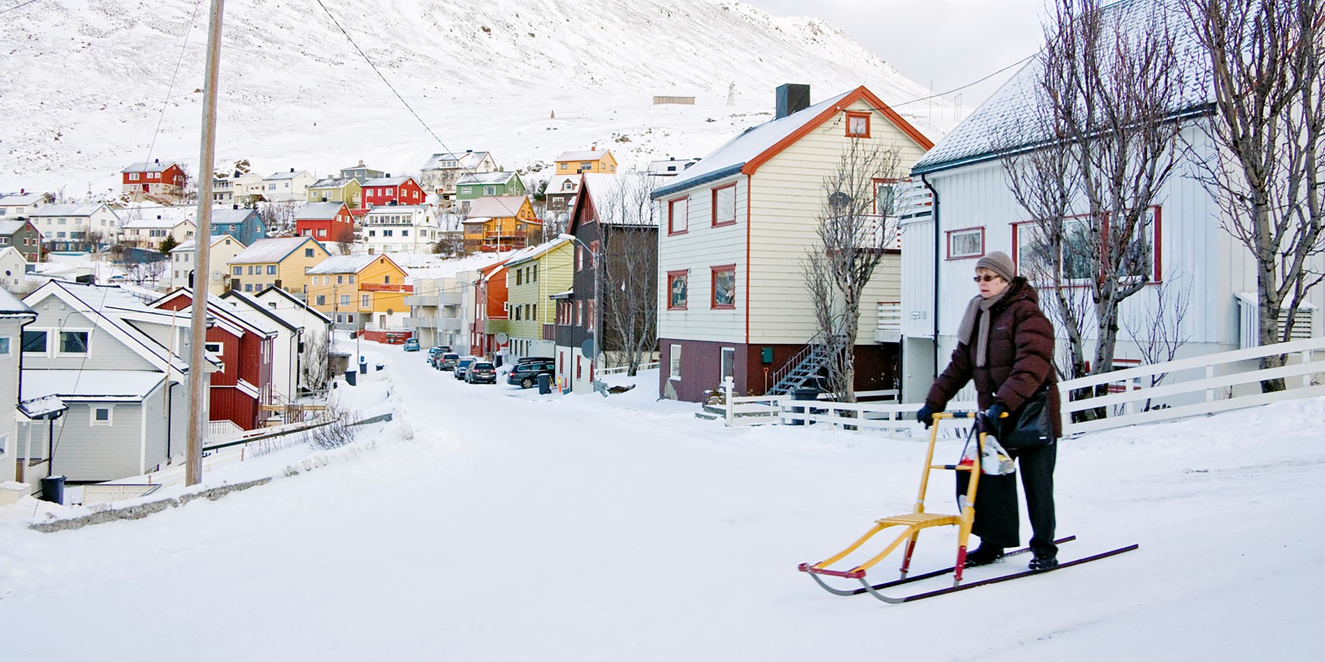 Et bilde av en eldre kvinne i snøen, med en gate med vakre, små og fargerike trehus i bakgrunnen