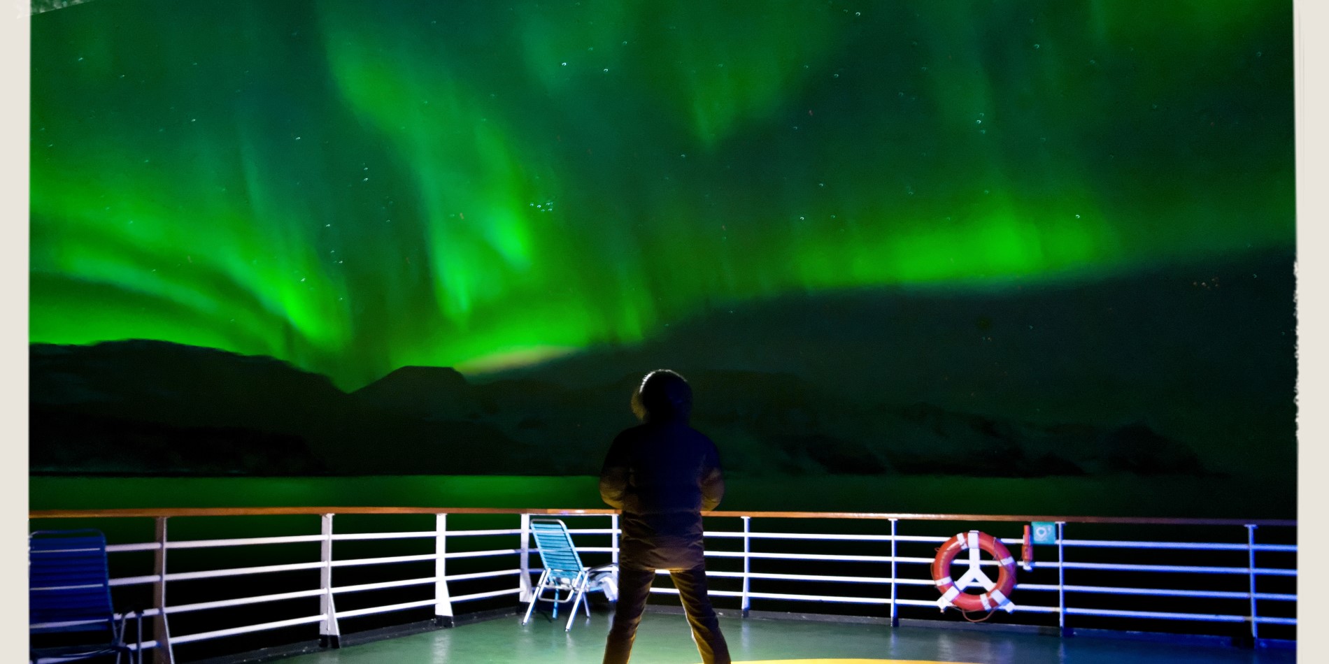 En mann står på dekk om bord på Hurtigruten på en reise langs norskekysten. Himmelen over ham lyser grønt – nordlyset danser