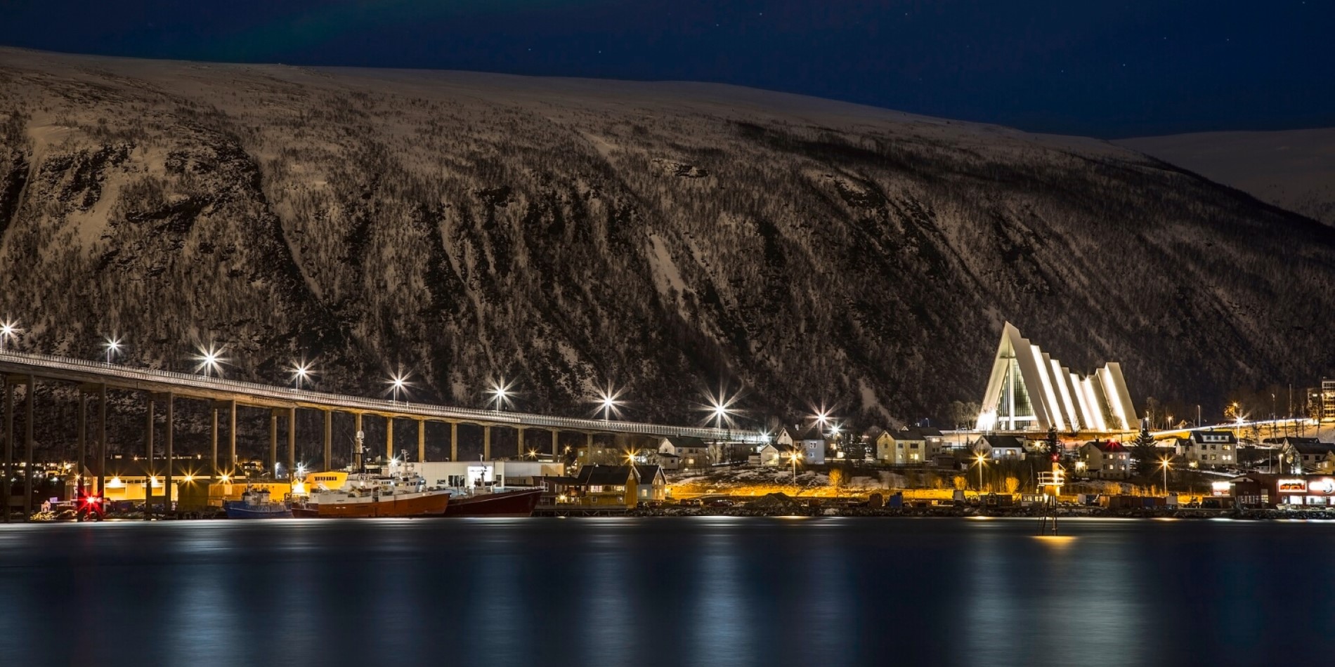 Ishavskatedralen sett fra Tromsøya. Broen og kirkebygget lyser opp den mørke kvelden